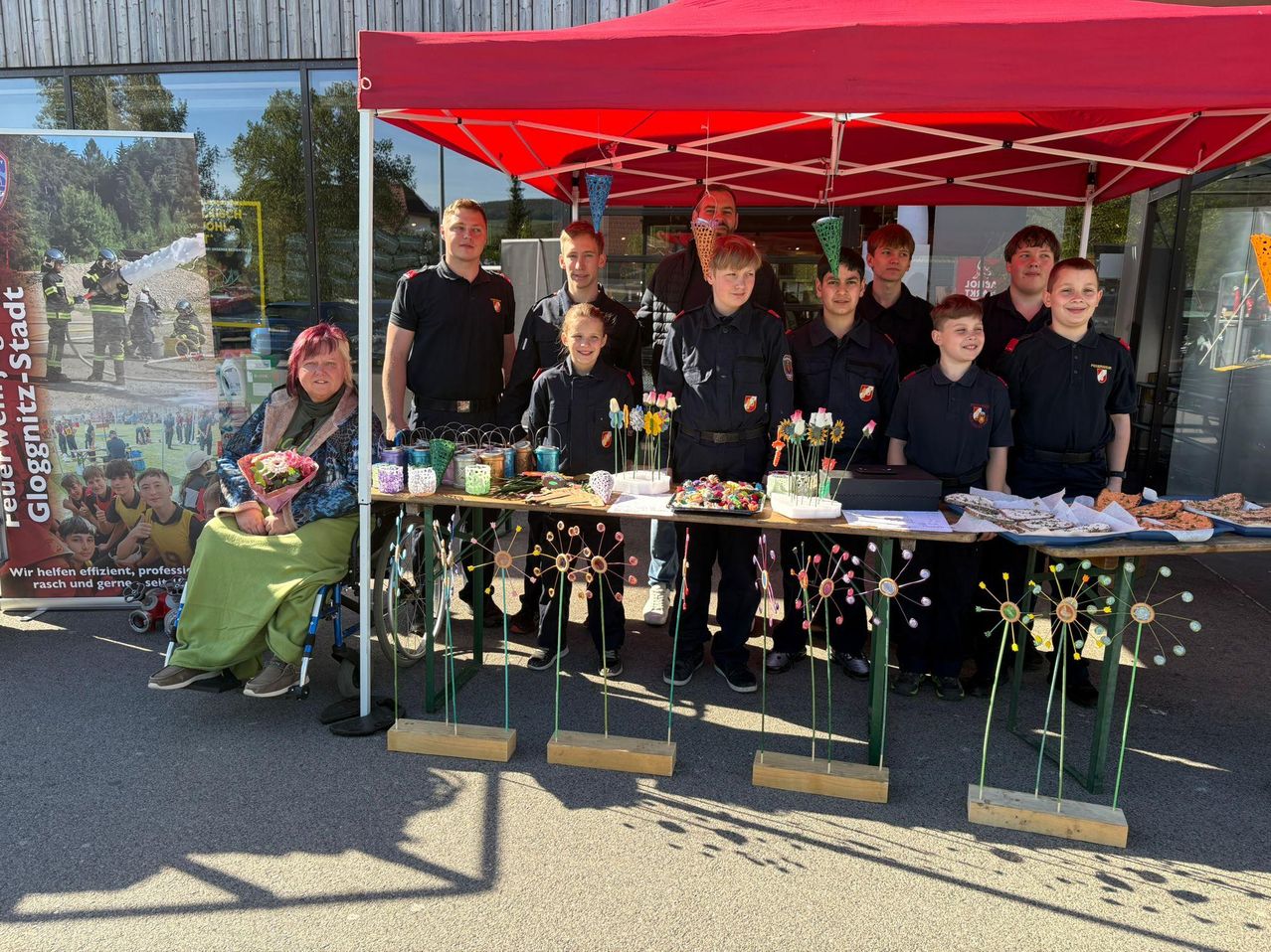 Eine Gruppe junger Leute in Uniform und eine Frau im Rollstuhl posieren für ein Foto unter einem Zelt mit verschiedenen ausgestellten Gegenständen.