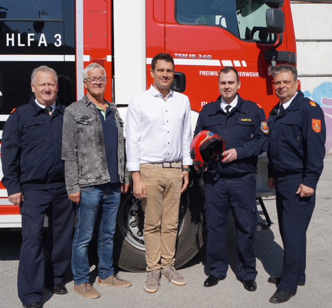 Five men in uniform stand in front of a fire truck labeled 'HLFA 3'. The central figure holds a red helmet.