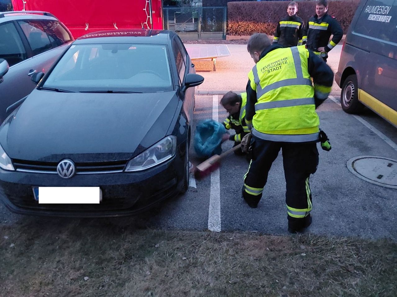Firefighters clean the road in front of a black Volkswagen car, two with brooms and two standing. A red fire truck is in the background.