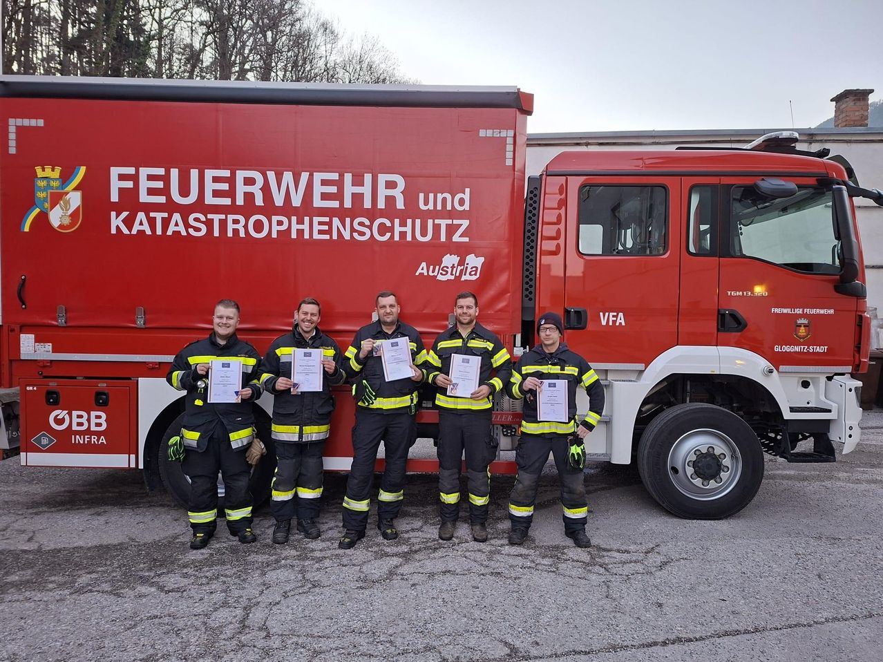 Fünf Feuerwehrleute stehen vor einem roten LKW mit der Aufschrift 'Feuerwehr und Katastrophenschutz'. Sie halten alle Urkunden in den Händen.