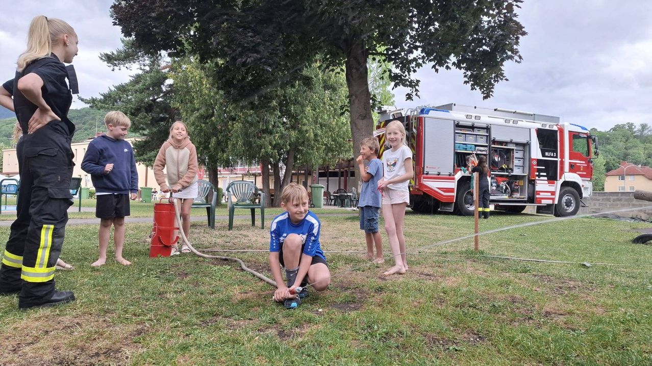Four children are playing with a water hose in a park. A fire truck is parked nearby.