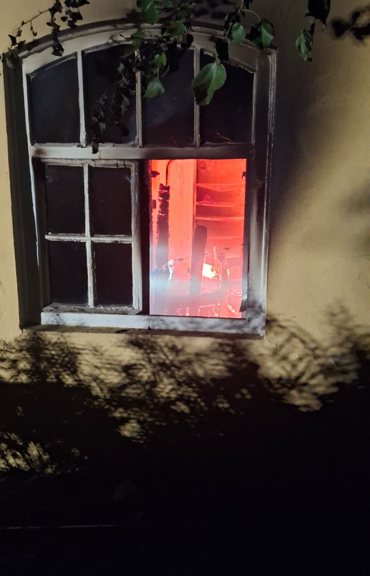 Ein Fenster mit einem Feuer im Inneren, das einen Raum mit Regalen und einem Feuerlöscher zeigt. Der Fensterrahmen ist beschädigt.