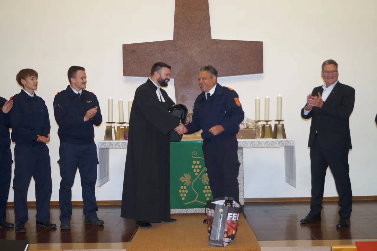 Ein Priester und ein Feuerwehrmann schütteln sich die Hände vor einem Altar mit Kerzen. Ein weiterer Feuerwehrmann und ein Mann stehen hinter ihnen.