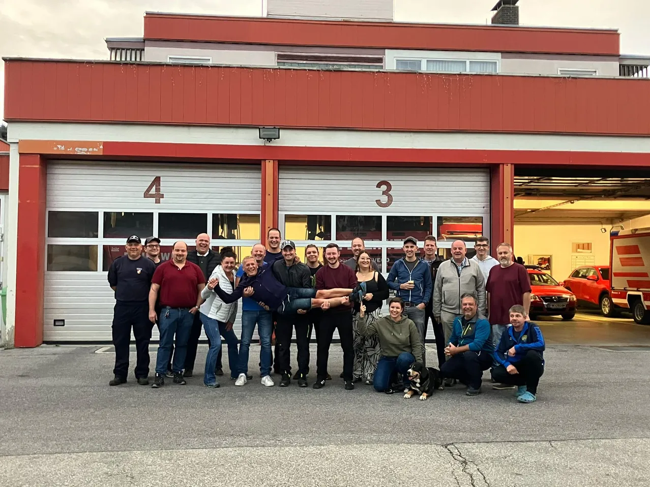 Eine Gruppe von Menschen, darunter Männer und Frauen, posiert für ein Foto vor einer Feuerwache. Die Feuerwache hat zwei Garagentore mit den Nummern 3 und 4.