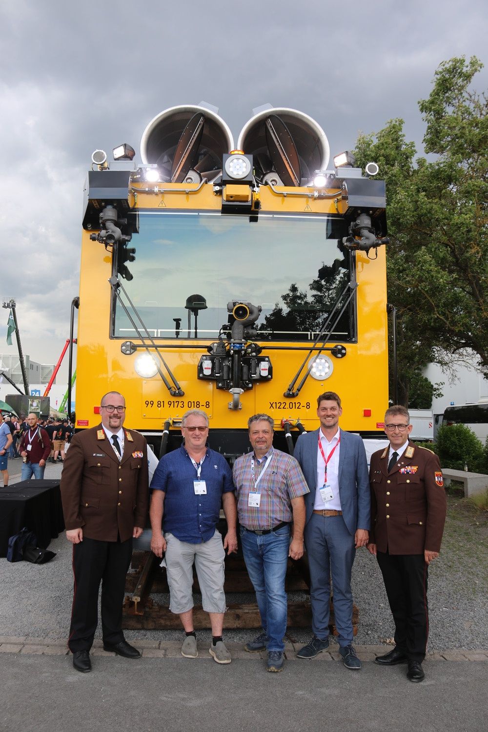Five men, four in suits, one in military attire, stand in front of a yellow locomotive with the number 99 81 9173 018-8. Behind them, there are people, trees, and buildings.