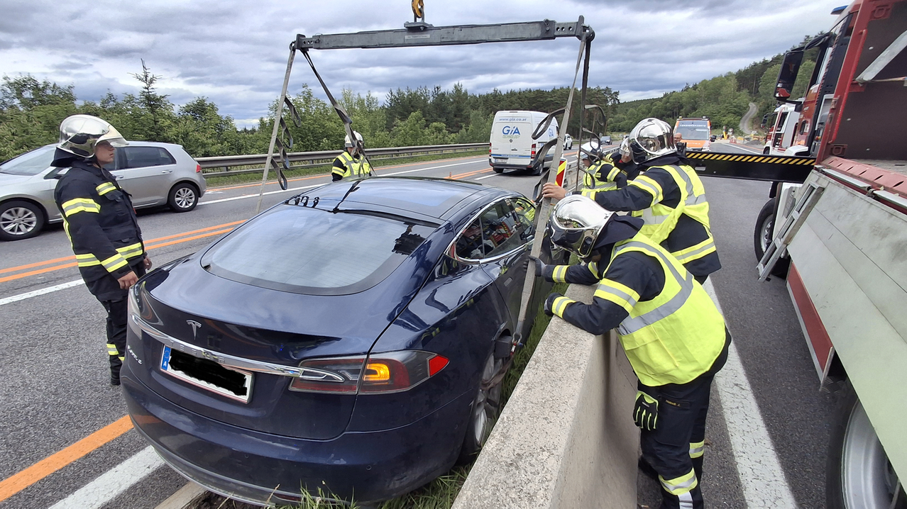 Rettungskräfte heben mit einem Kran einen blauen Tesla Model 3 von der Straße. Das Auto ruht auf dem Gras neben der Betonbarriere. Sie tragen Helme und Warnwesten.