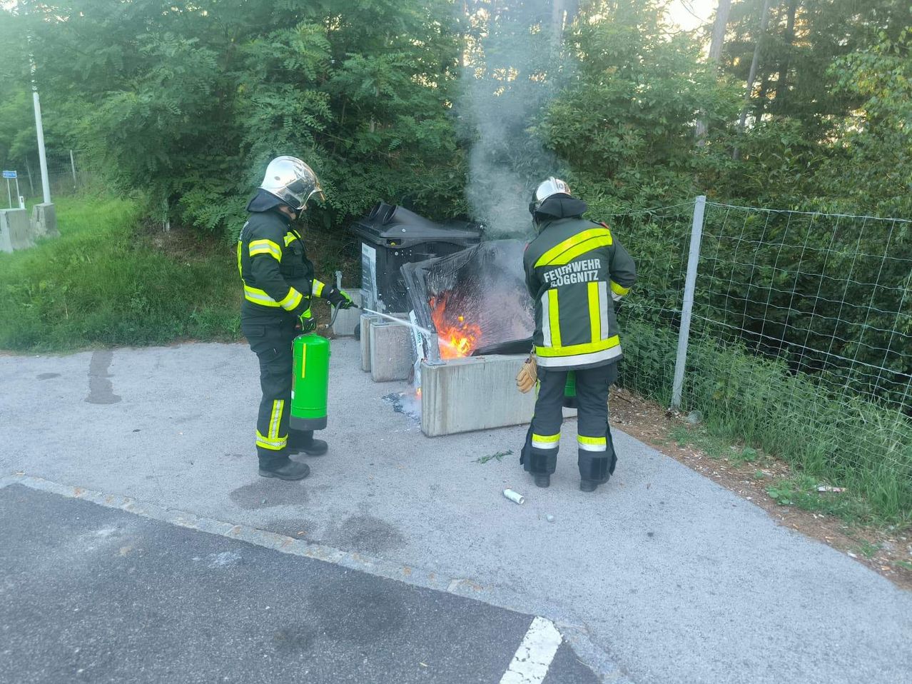 Zwei Feuerwehrleute stehen neben einem brennenden Container, einer hält eine Wasserspritze und der andere benutzt einen Feuerlöscher. Sie befinden sich auf einer Betonstraße mit viel Grün und einem Zaun im Hintergrund.