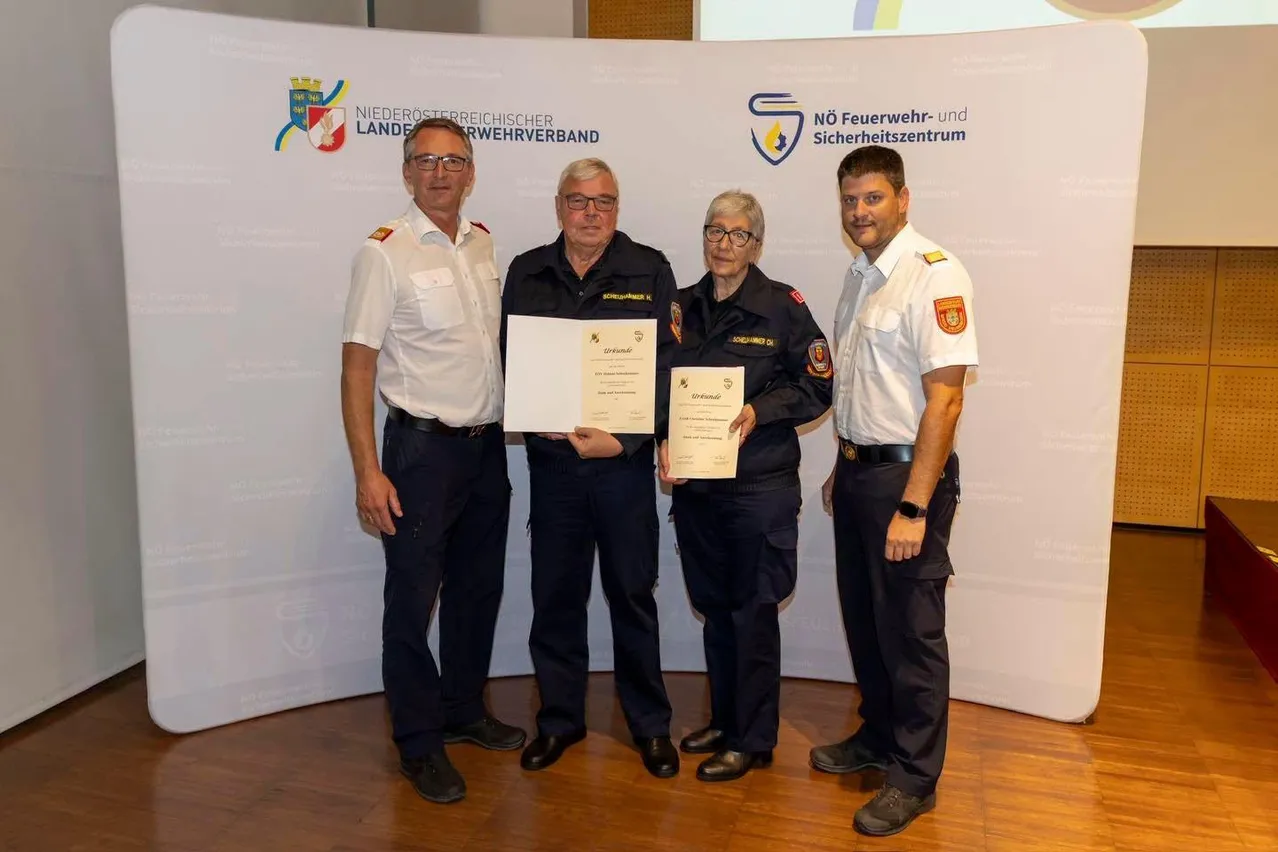 Vier Personen in Uniform stehen zusammen und halten Urkunden in der Hand. Sie lächeln und scheinen bei einer Preisverleihung zu sein. Hinter ihnen ist ein Hintergrund mit Logos und Text.