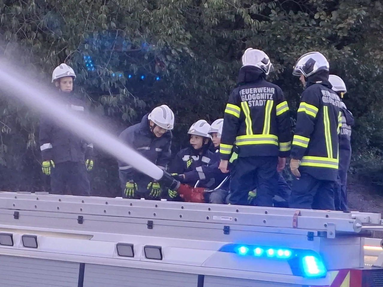 Feuerwehrleute in Schutzausrüstung, einschließlich Helmen und Handschuhen, sprühen Wasser aus einem Feuerwehrauto. Mehrere Kinder in Helmen beobachten.