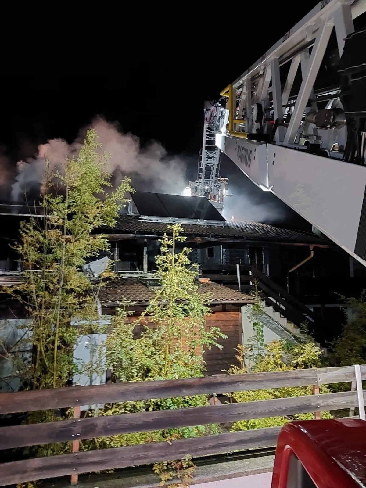 Ein Feuerwehrfahrzeug steht bei Nacht neben einem brennenden Gebäude. Rauch steigt vom Dach auf, und Feuerwehrleute sind auf dem Dach sichtbar.