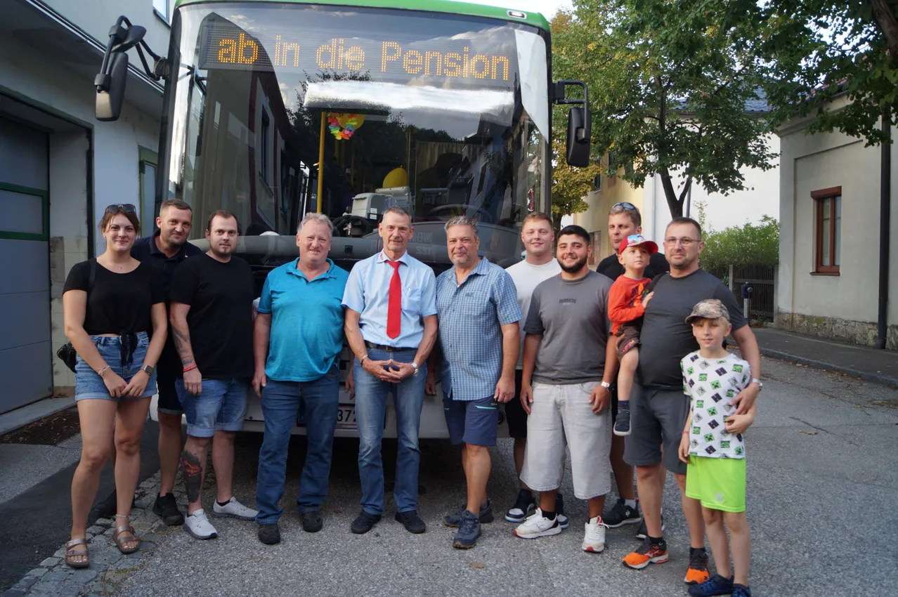 Eine Gruppe von Menschen steht vor einem Bus mit einer digitalen Anzeige, die 'ab in die Pension' anzeigt.