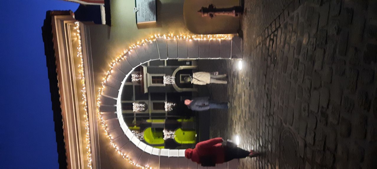 Zwei Personen gehen unter einem dekorativen Bogen mit Lichterketten. Der Bogen hat einen Spiegel, der ein grünes Objekt reflektiert. Ein Feuerhydrant befindet sich auf der rechten Seite der Wand.