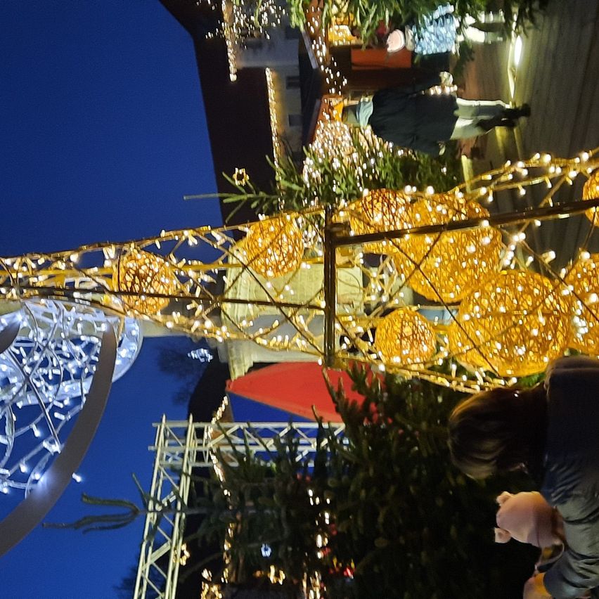 Ein mit gelben Lichtern und Ornamenten geschmückter Weihnachtsbaum steht in einem festlichen Bereich. Menschen, darunter eine Frau mit einem Baby, gehen herum.