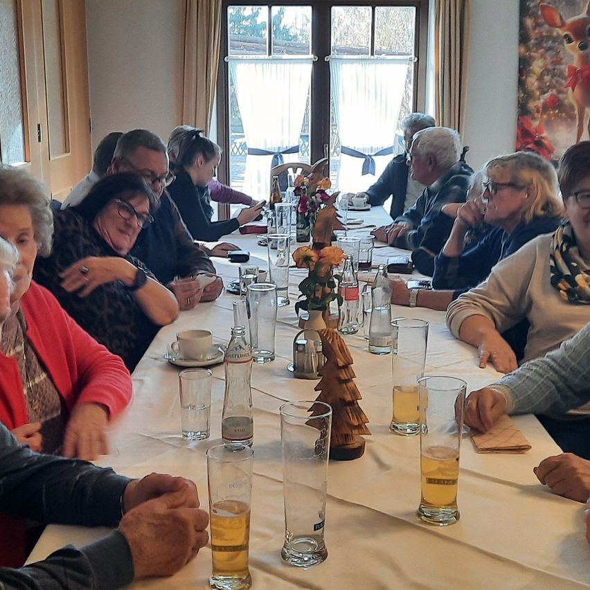 Eine Gruppe von Menschen, einige mit Brille, sitzt um einen Tisch mit Getränken und Essen. Der Tisch ist mit Gläsern, einer Blumenvase und anderen Gegenständen gedeckt. Der Raum hat Fenster mit Vorhängen und ein Bild an der Wand.