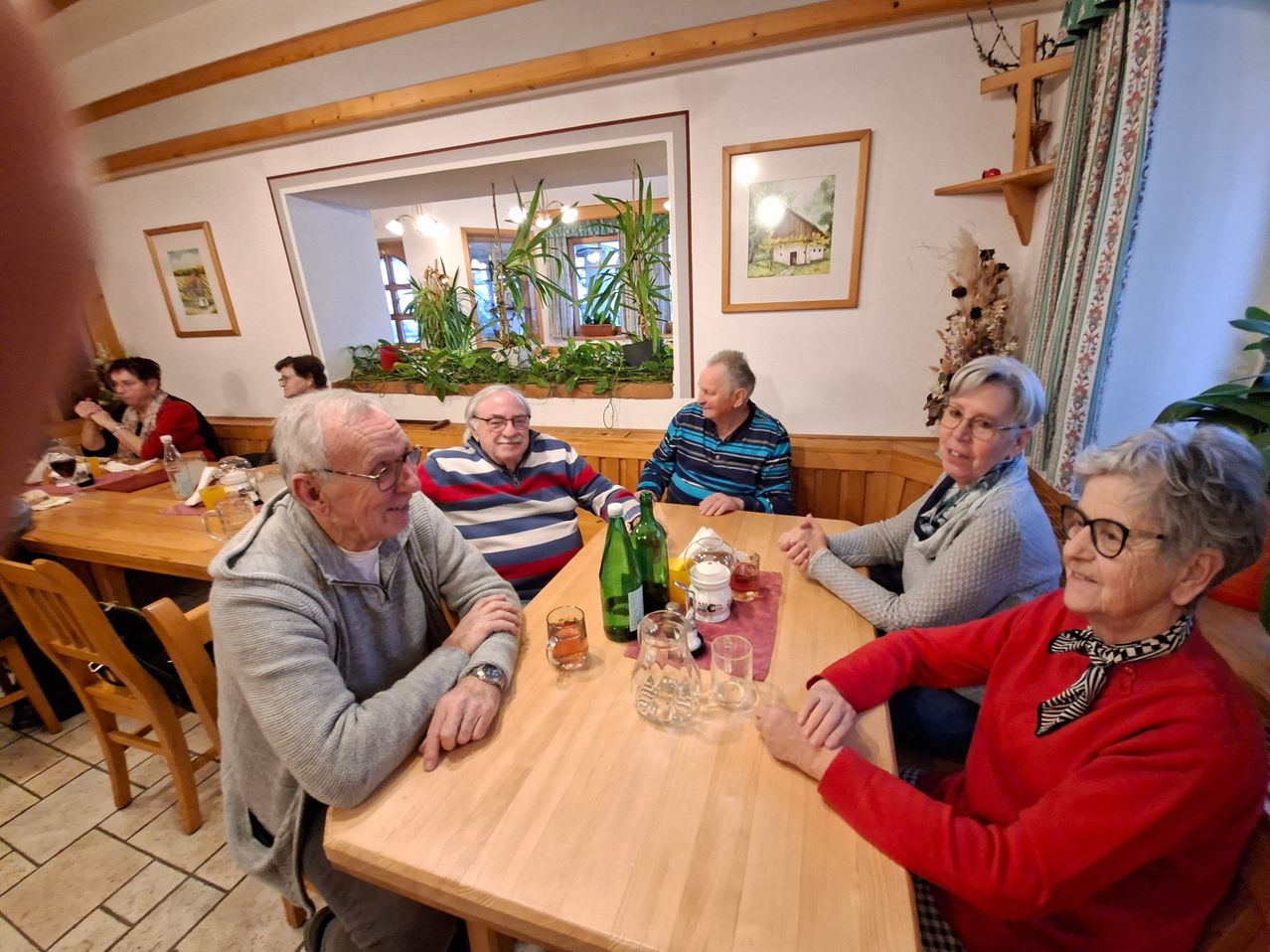 A group of elderly people are sitting around a wooden table in a restaurant. They are smiling and seem to be enjoying their conversation. There are bottles, glasses, and other items on the table. The room has wooden walls and a window with a view of a garden.