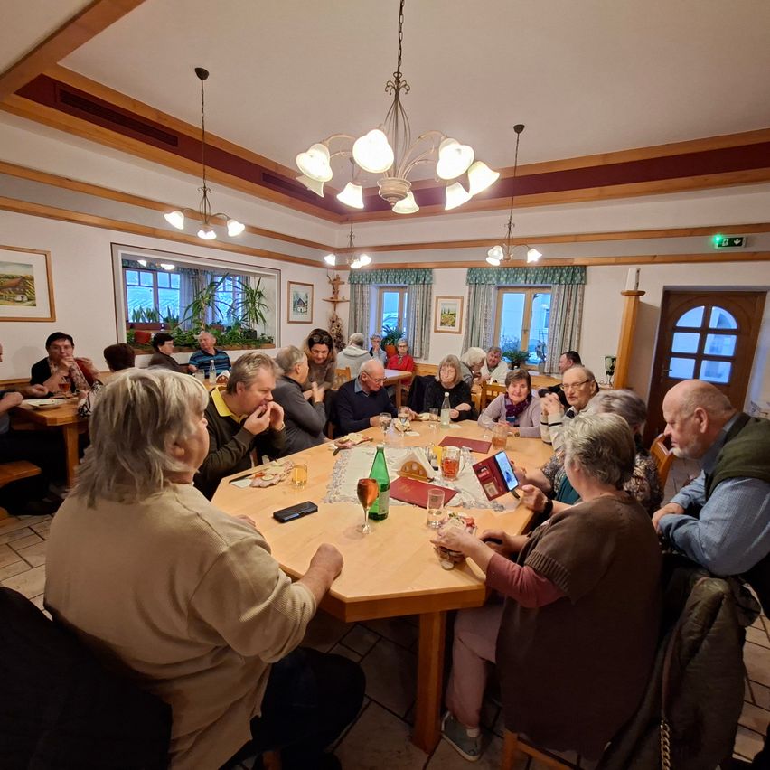 A group of people sit around a table in a dining room. A few people are eating, and others are talking. There are bottles, glasses, and plates on the table.