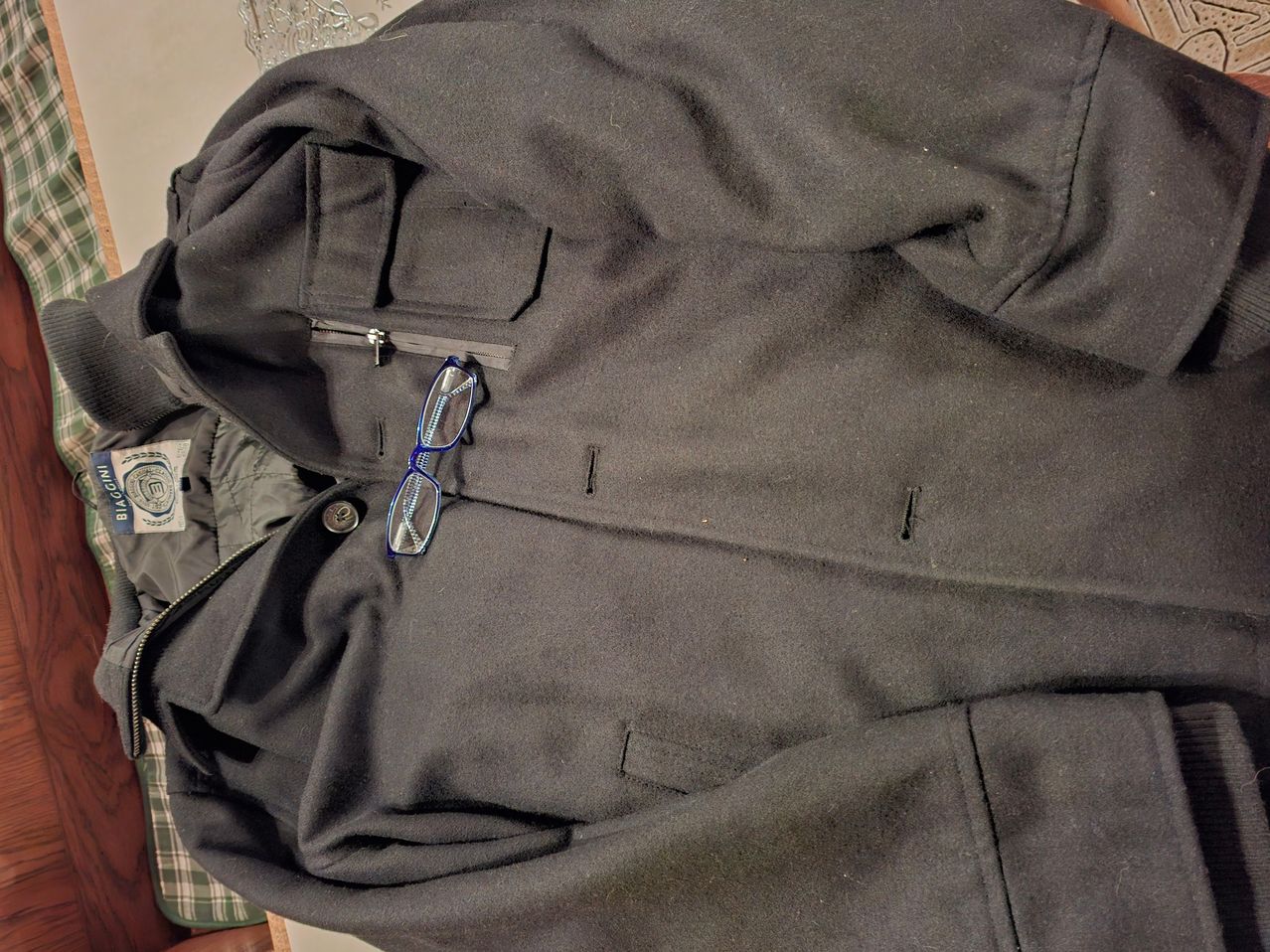 Ein schwarzer Mantel mit Reißverschluss und Taschen ist gefaltet, und eine Brille in Blau liegt darauf.