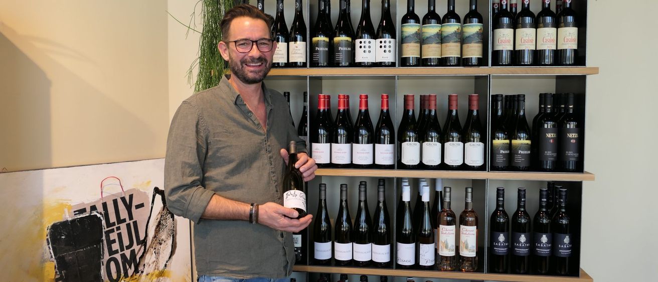 Ein Mann steht vor einem Holzregal voller verschiedener Weinflaschen. Er hält eine Flasche in der Hand und lächelt.