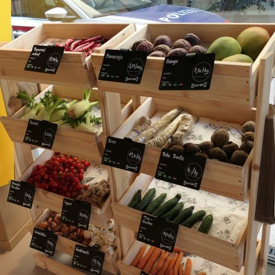 Ein Holzregal zeigt verschiedene Gemüse und Früchte in verschiedenen Fächern, darunter Paprika, Fenchel, Radieschen, Karotten und Gurken, mit Preisschildern.