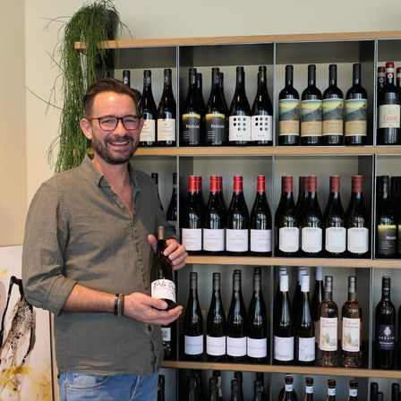 Ein Mann lächelt, während er eine Weinflasche vor einem Weinregal voller verschiedener Weine hält.