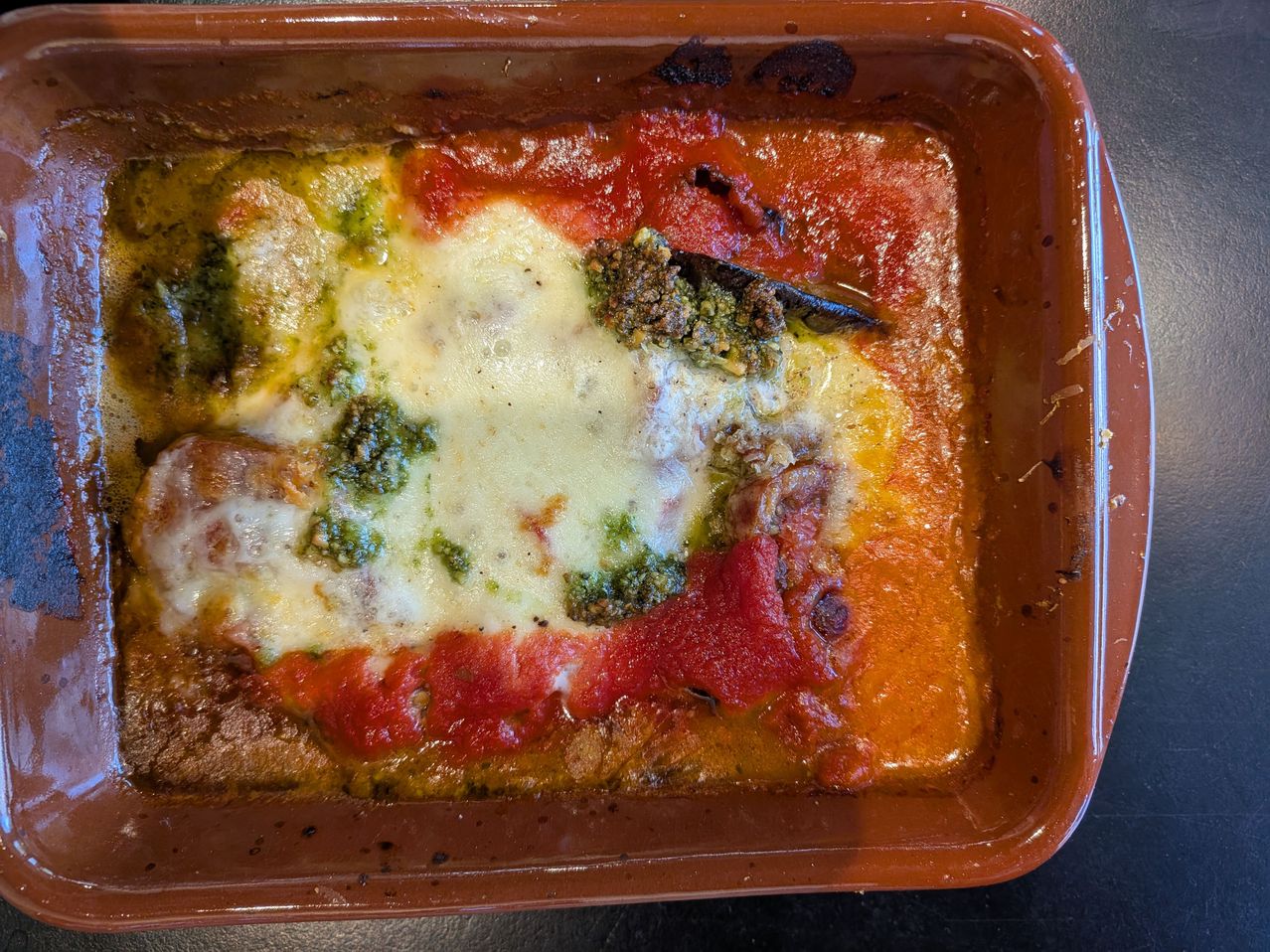A close-up of a baked dish in a brown tray. It has layers of pasta, red sauce, green pesto, and melted cheese. The dish is slightly burnt on the edges.