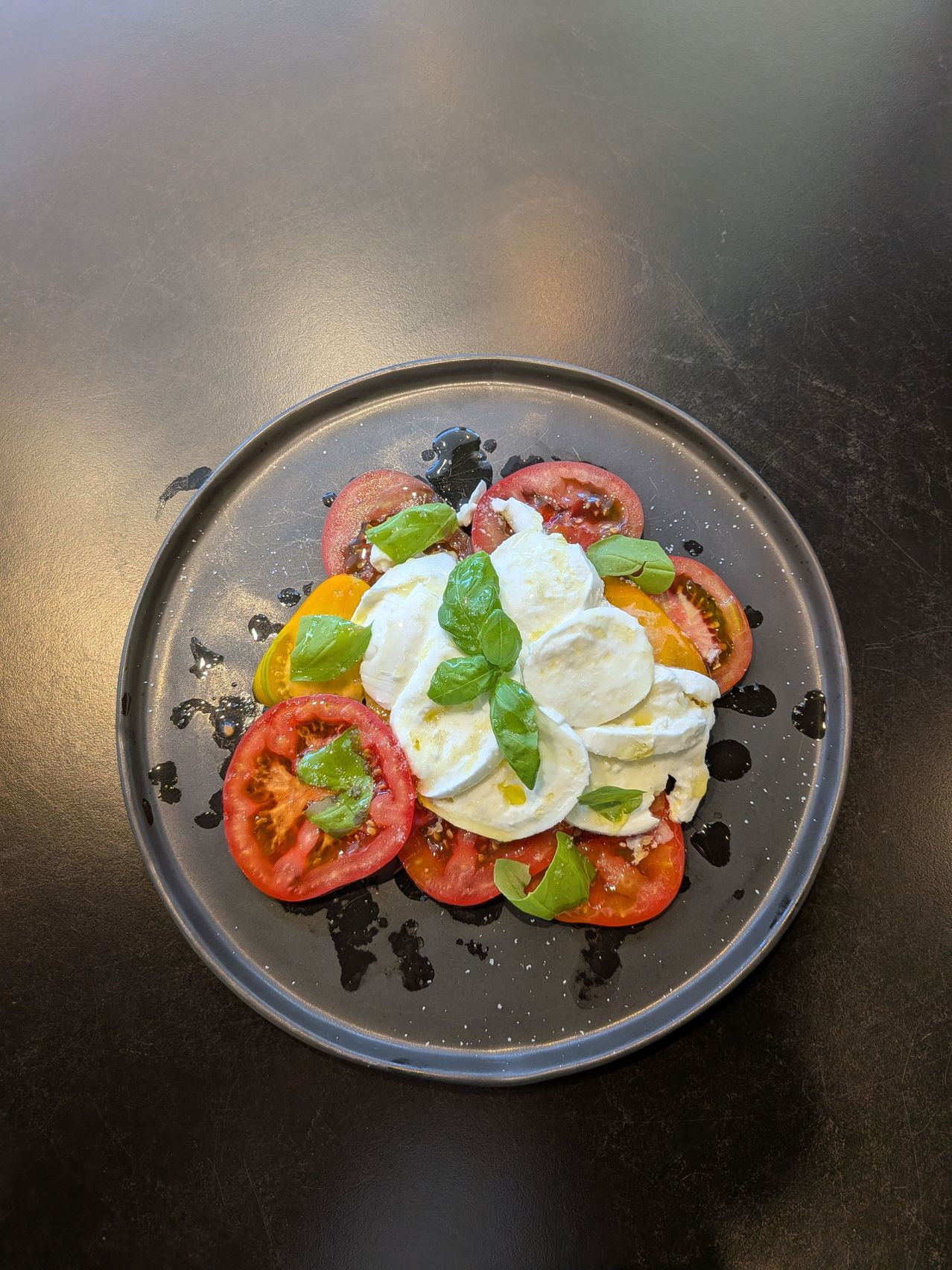 Ein Teller mit einem frischen Salat aus Tomatenscheiben, Mozzarella und Basilikumblättern auf einer dunklen Oberfläche.