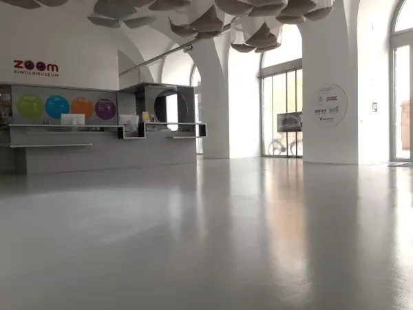 Ein leerer Empfangsbereich mit einem weißen Schreibtisch, Monitoren und farbigen Kreisen an der Wand. Darüber befindet sich ein einzigartiges Deckendesign mit hängenden Formen und großen Glasfenstern.