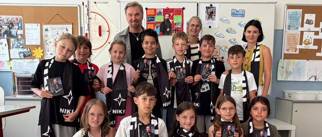 Eine Gruppe von Kindern und Erwachsenen in einem Klassenzimmer. Sie alle lächeln und posieren für ein Foto. Sie tragen schwarze und weiße Schals mit dem Namen Niki.
