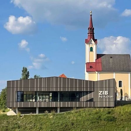 Eine Kirche mit einem Kirchturm steht prominent in einem Dorf, dahinter ein modernes Gebäude, umgeben von Häusern, Bäumen und einem Feld unter einem blauen Himmel mit verstreuten Wolken.