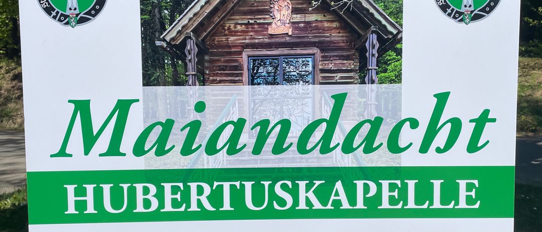 Ein Schild für Maiandacht Hubertuskappele am Freitag, 01. Mai, um 18 Uhr, mit einem Bild eines Holzhauses.