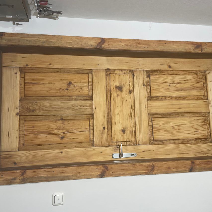 Eine Holztür mit einem silbernen Griff ist an einer weißen Wand montiert. Die Tür hat mehrere Paneele mit sichtbaren Holzmaserungen. Ein Metallobjekt ist am unteren Teil der Tür befestigt.