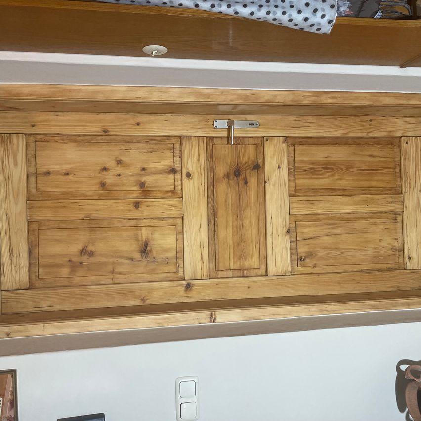 Ein hölzerner Schrank mit drei Türen, wobei die mittlere Tür mit einem Metallverschluss versehen ist. Oben befindet sich ein hölzernes Regal mit darauf liegenden Gegenständen. Unten ein weißer Wand mit einem Schalter.