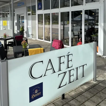 Der Eingang eines Cafes mit Glastüren und Tischen mit Stühlen und Blumen draußen. Der Name des Cafés ist Cafe Zeit.