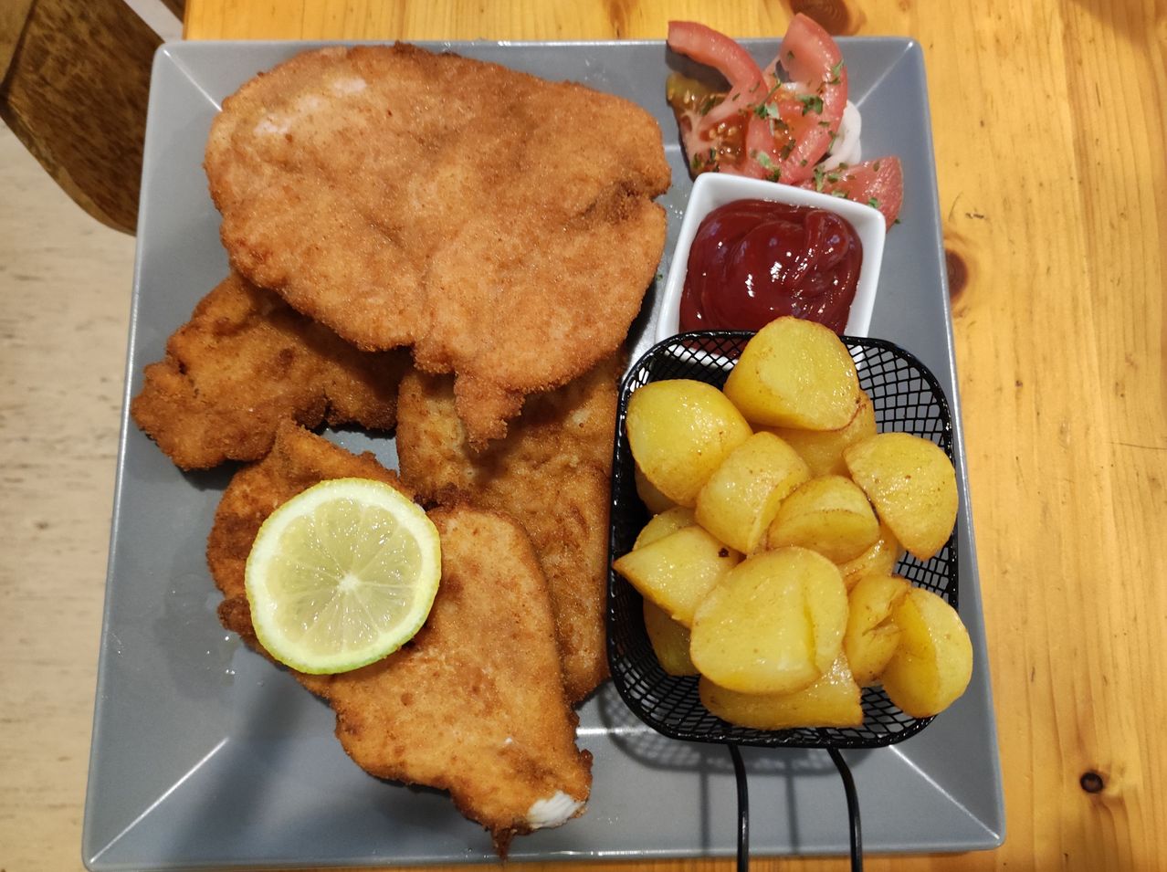Ein Teller mit gebratenem Fisch, Zitronenscheibe, Tomaten und Ketchup. Daneben sind gekochte Kartoffeln in einem Korb.