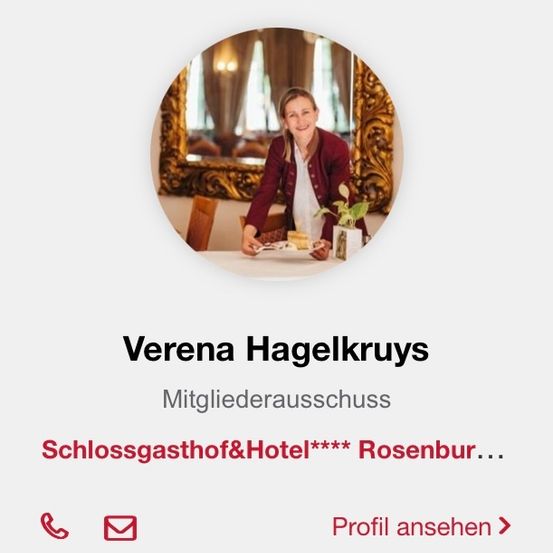 Eine lächelnde Frau steht vor einem Tisch, hält einen Teller mit Essen und eine Topfpflanze in der Hand. Sie trägt eine burgunderfarbene Strickjacke über einem weißen Hemd. Dahinter befindet sich ein Spiegel mit einem kunstvollen Rahmen. Unten gibt es einen Aufruf zum Ansehen des Profils.