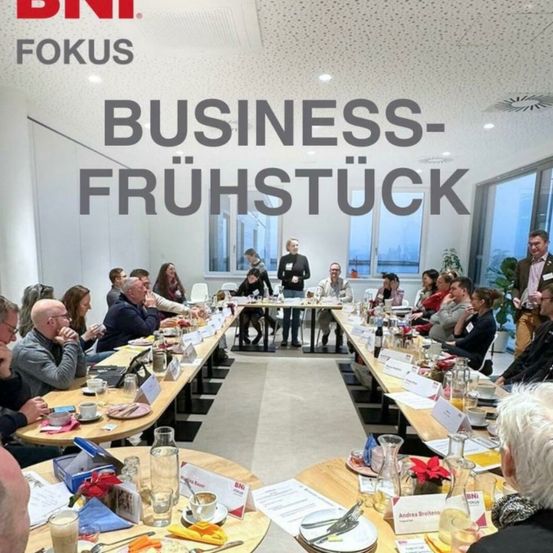 Businessfruhstuck-Event mit einer großen Gruppe von Menschen, die an Tischen mit Essen und Getränken sitzen, eine Person steht mit einem Mikrofon an der Spitze des Tisches.