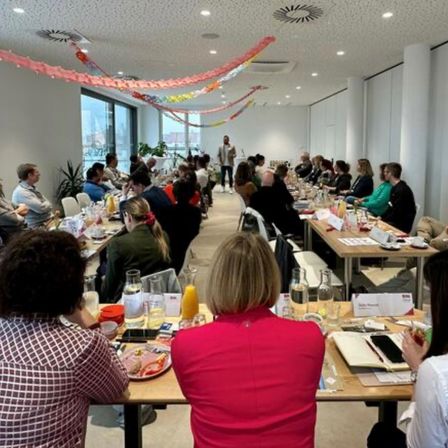 Eine Gruppe von Menschen sitzt an einem langen Tisch in einem Konferenzraum. Der Raum ist mit bunten Girlanden dekoriert, und es gibt Pflanzen und Glasobjekte auf dem Tisch. Ein Mann steht vor ihnen und hält wahrscheinlich eine Präsentation.