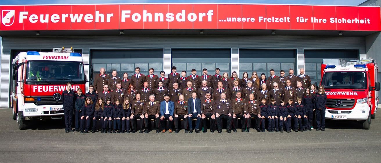 Eine Gruppe uniformierter Personen, einschließlich Männer, Frauen und Kinder, steht vor einem Gebäude mit einem roten Schild, auf dem 'ehr Fohnsdorf...unsere Freiheit' steht. Sie posieren für ein Gruppenfoto.