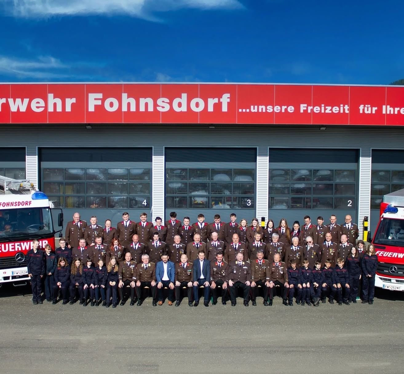 Eine Gruppe uniformierter Personen, möglicherweise Feuerwehrleute, posiert für ein Foto vor einer Feuerwache. Das Gebäude hat ein rotes Schild mit der Aufschrift 'wehr Fohnsdorf'. Vor dem Gebäude sind Feuerwehrwagen geparkt.