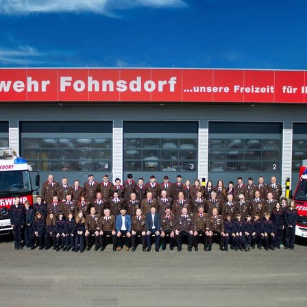 Eine Gruppe uniformierter Personen, möglicherweise Feuerwehrleute, posiert für ein Foto vor einer Feuerwache. Das Gebäude hat ein rotes Schild mit der Aufschrift 'wehr Fohnsdorf'. Vor dem Gebäude sind Feuerwehrwagen geparkt.