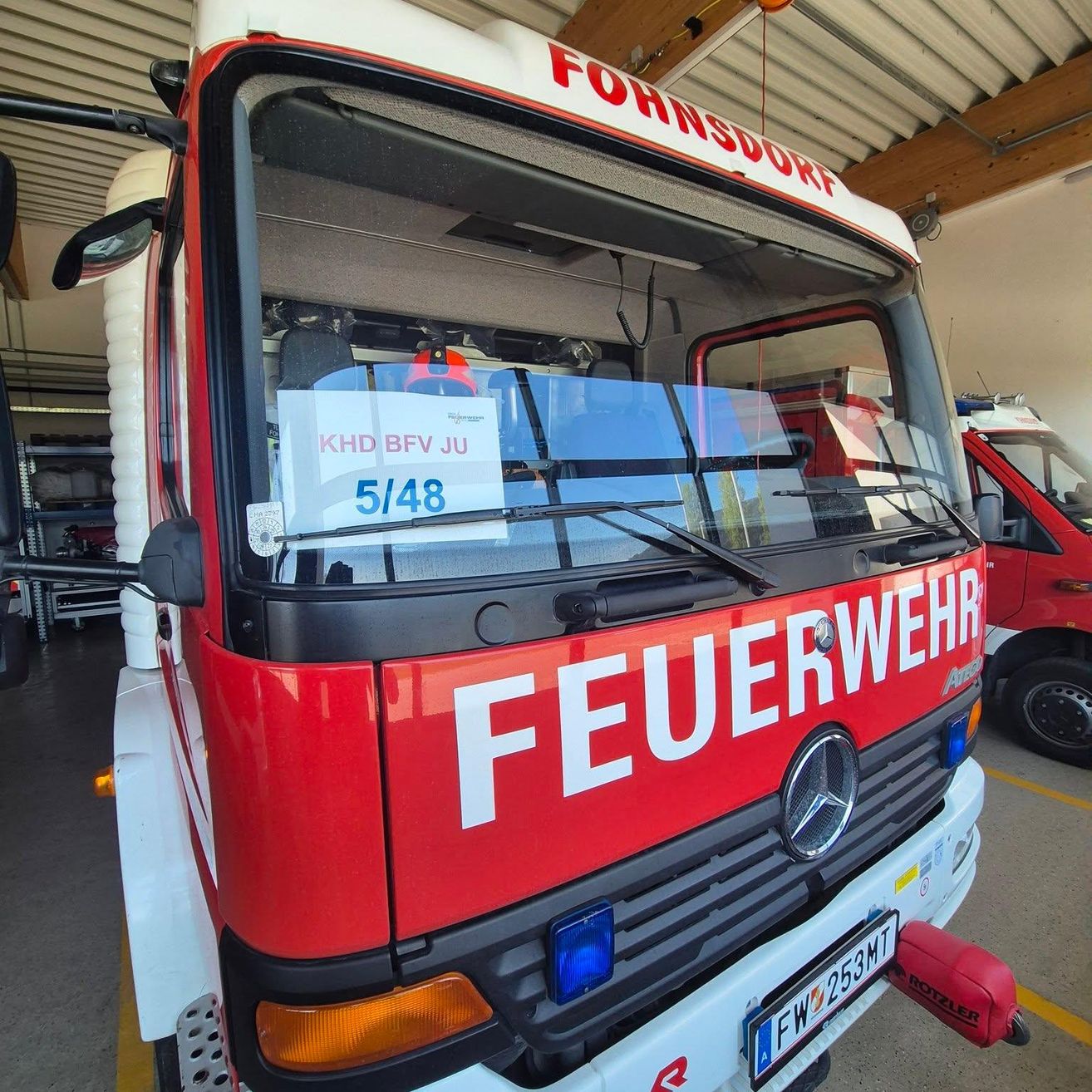 Ein roter und weißer Feuerwehrwagen mit dem Wort 'Feuerwehr' auf der Vorderseite, in einer Garage geparkt. Der Wagen hat ein Nummernschild mit der Nummer 'FW 253MT'.