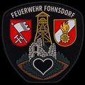 Ein Feuerwehrabzeichen mit dem Namen 'Feuerwehr Fohnsdorf' oben, mit einem Turm, einem Herz und zwei Wappen auf beiden Seiten. Der Hintergrund ist schwarz.