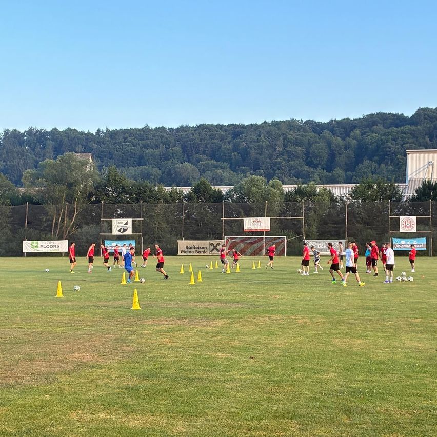 Eine Fußballtrainingseinheit findet im Freien statt, bei der Spieler in roten und blauen Trikots trainieren. Sie befinden sich zwischen Hütchen auf einem Feld mit einem Netz in der Mitte. Im Hintergrund sind Werbebanner zu sehen.