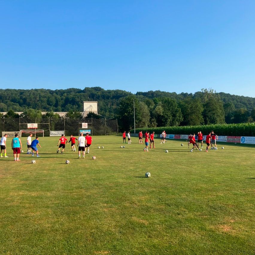 Eine Gruppe von Menschen in roten und weißen Trikots übt Fußball auf einem Rasenfeld mit Fußballbällen. Der Himmel ist blau, und die Umgebung ist mit Bäumen und einem Basketballkorb gefüllt.
