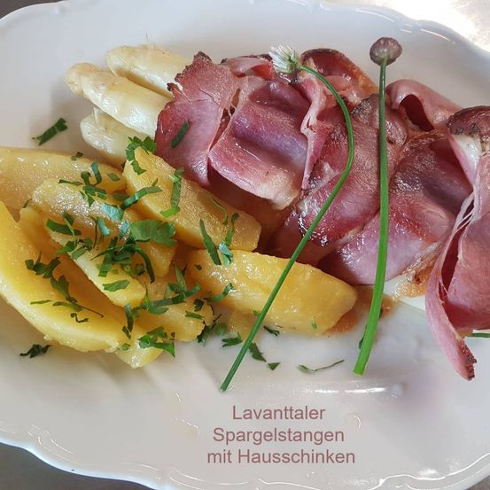 Ein Teller mit geschnittenen Kartoffeln, Spargeln und Hausschinken, garniert mit Kräutern, wird mit dem Text 'Lavanttaler Spargelstangen mit Hausschinken' präsentiert.