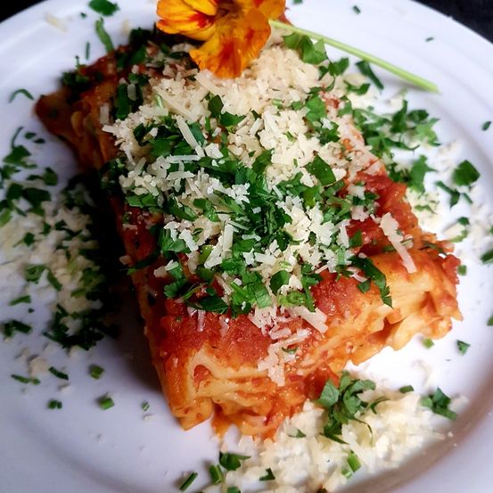 Ein Gericht von Lasagne auf einem Teller, garniert mit geriebener Käse, Kräutern und einer gelben Blume.