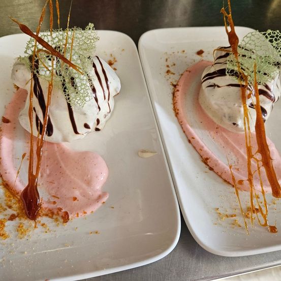 Zwei weiße Teller mit Desserts, jeweils mit einer Baiserhaube und Schokoladensauce garniert, sowie einer rosafarbenen Soße. Die Baiserhauben haben braune Streifen und sind mit orangefarbenen Fäden verziert.