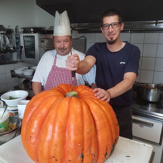 Zwei Männer in einer Küche, einer hält ein Messer und will einen großen Kürbis schneiden. Hinter ihnen befinden sich Küchengeräte und Utensilien.