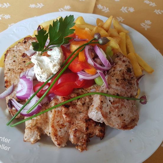Ein Teller mit gegrilltem Hähnchen, Pommes Frites und einer Mischung aus Gemüse, darunter Tomaten und Zwiebeln. Das Gericht ist mit Petersilie garniert und auf einem weißen Teller serviert.