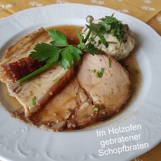 Ein weißer Teller mit geschnittenem Schweinebraten, garniert mit Petersilie, serviert mit Kartoffelpüree, alles in einer Holzschüssel.
